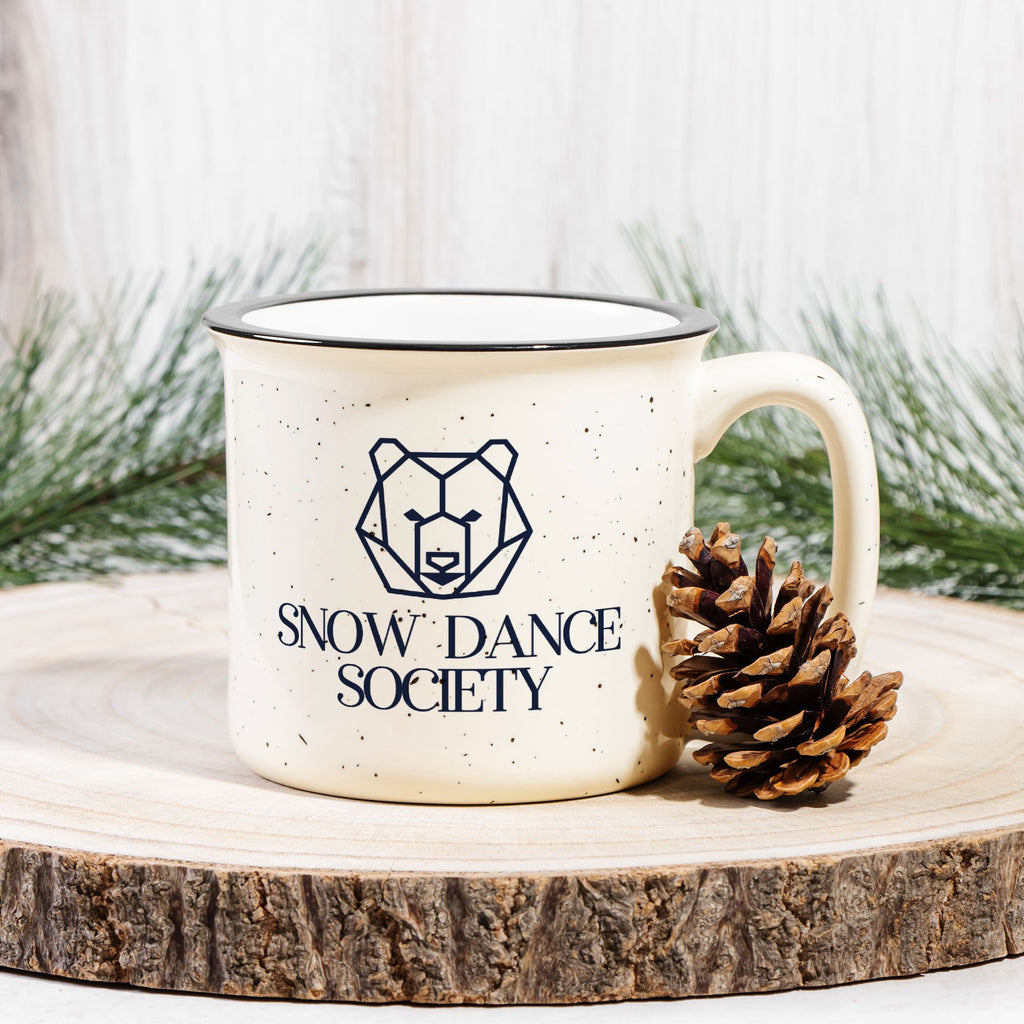 Snow Dance Society camp mug styled with pine branch and pinecone on a wood slab