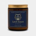 Larch Season candle in an amber glass jar with a navy and gold Snow Dance Society label